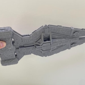 Op de afbeelding: Een grijs, gedetailleerd model van een ruimteschip, vastgehouden door een hand. Het model heeft een complex ontwerp met verschillende panelen en structuren, die lijken op een futuristisch ruimteschip. De kleur is een uniform grijs.