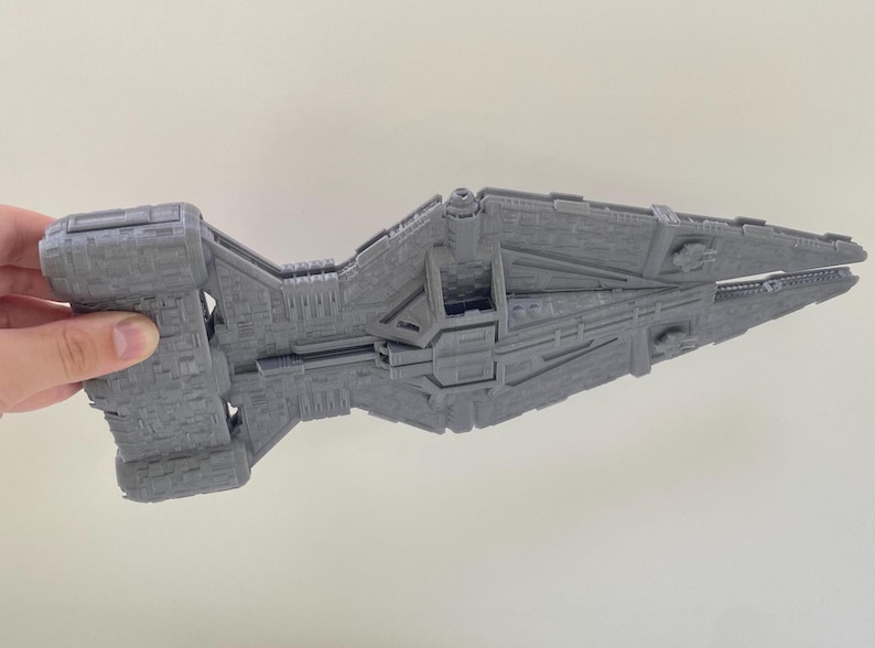 Op de afbeelding: Een grijs, gedetailleerd model van een ruimteschip, vastgehouden door een hand. Het model heeft een complex ontwerp met verschillende panelen en structuren, die lijken op een futuristisch ruimteschip. De kleur is een uniform grijs.