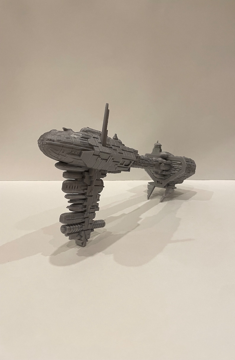 May include: A grey 3D printed model of a spaceship with a long, thin body and a large, round cockpit. The spaceship has a series of small, rectangular windows along the side of the body.