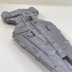 Op de afbeelding: Een gedetailleerd, grijs model van een ruimteschip met ingewikkelde panelen en motordetails. Het model heeft een textuuroppervlak en een symmetrisch ontwerp, wat wijst op een sciencefictionthema. Het ontwerp van het schip omvat meerdere rechthoekige en cilindrische componenten.