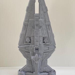 Op de afbeelding: Grijs 3D-geprint model van een ruimteschip met ingewikkelde details. Het model heeft een centraal lichaam met een gespleten bovenkant, gedetailleerde panelen en cilindrische basissteunen. Het ontwerp suggereert een futuristisch of sciencefiction thema.