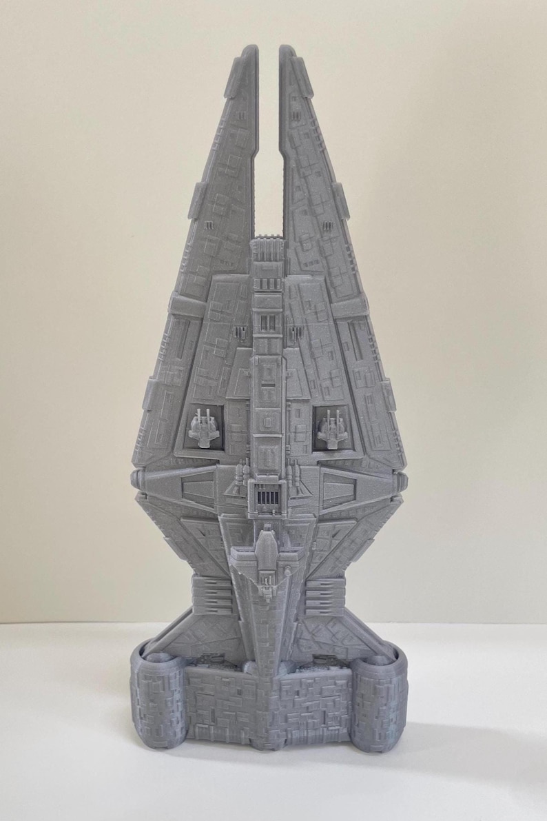 Op de afbeelding: Grijs 3D-geprint model van een ruimteschip met ingewikkelde details. Het model heeft een centraal lichaam met een gespleten bovenkant, gedetailleerde panelen en cilindrische basissteunen. Het ontwerp suggereert een futuristisch of sciencefiction thema.