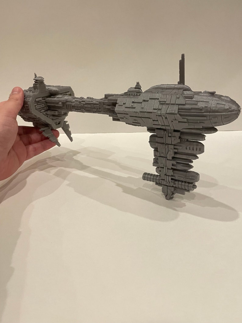 May include: A grey 3D printed model of a spaceship with a long, thin body and a large, round engine section. The model is detailed with many small parts and features.
