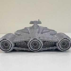 Op de afbeelding: Een grijs, 3D-geprint model van een ruimteschip met drie grote motorstraalpijpen en ingewikkelde details. Het ruimteschip heeft een centraal lichaam met een verhoogd gedeelte en meerdere kleinere cirkelvormige kenmerken.