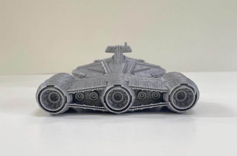Op de afbeelding: Een grijs, 3D-geprint model van een ruimteschip met drie grote motorstraalpijpen en ingewikkelde details. Het ruimteschip heeft een centraal lichaam met een verhoogd gedeelte en meerdere kleinere cirkelvormige kenmerken.