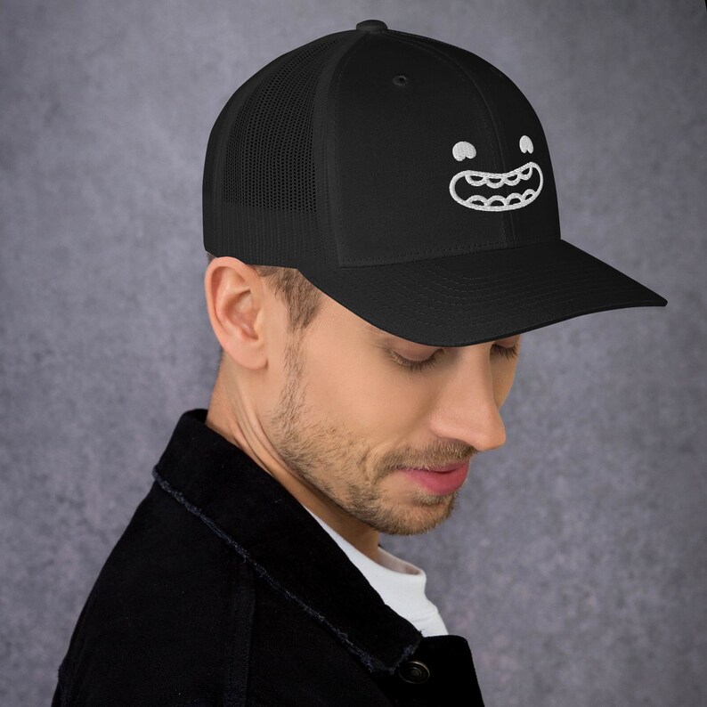 Puede incluir: Una gorra de camionero negra con una cara de dibujos animados bordada en blanco en la parte delantera. La cara tiene una amplia sonrisa con dientes y dos ojos grandes.