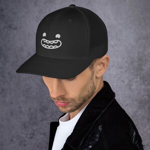 Puede incluir: Una gorra de camionero de malla negra con una cara sonriente bordada en blanco con dientes. La gorra tiene una visera negra y una parte trasera de malla negra.