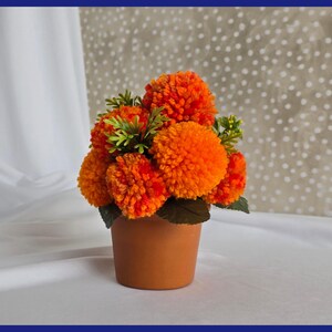 May include: A terracotta pot filled with a bouquet of orange and yellow yarn pom-poms, resembling a flower arrangement.