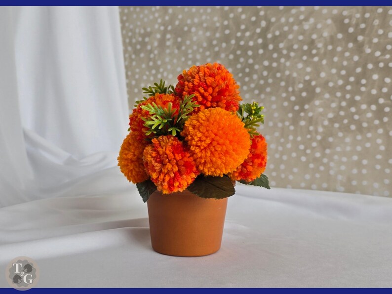 May include: A terracotta pot filled with a bouquet of orange and yellow yarn pom-poms, resembling a flower arrangement.