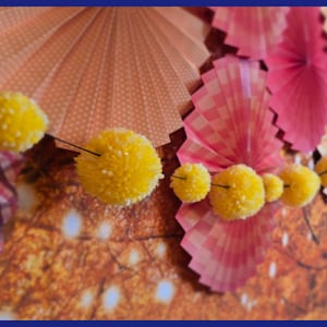 May include: A garland of yellow pom-poms hangs in front of a backdrop of pink and orange paper fans. The fans have a checkered pattern and are folded in a fan shape.