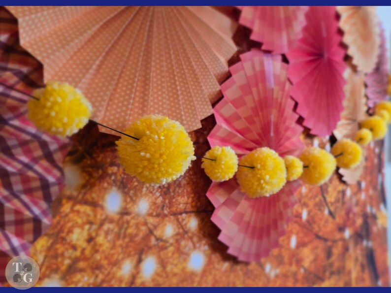 May include: A garland of yellow pom-poms hangs in front of a backdrop of pink and orange paper fans. The fans have a checkered pattern and are folded in a fan shape.