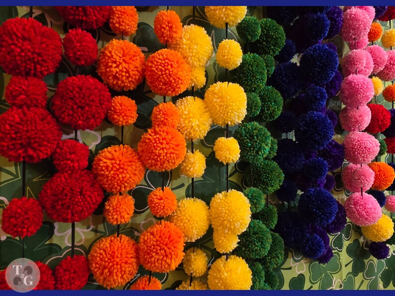 May include: A wall decorated with colorful pom-poms in a rainbow pattern. The pom-poms are made of yarn and are strung together on a string. The background is a green and white pattern.