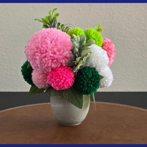May include: A decorative floral arrangement featuring a variety of colorful pom-poms in shades of pink, green, and white. The pom-poms are arranged with faux greenery in a small, off-white ceramic vase, creating a cheerful display.