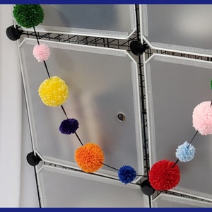 May include: A colorful pom-pom garland with alternating colors of pink, yellow, green, blue, orange, red, and purple. The garland is hanging from a white wire rack.