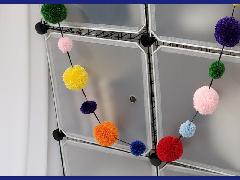 May include: A colorful pom-pom garland with alternating colors of pink, yellow, green, blue, orange, red, and purple. The garland is hanging from a white wire rack.