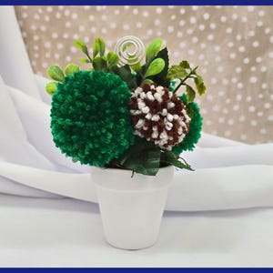 May include: A small white pot with green and brown yarn pom-poms, resembling flowers, in a green leafy arrangement.