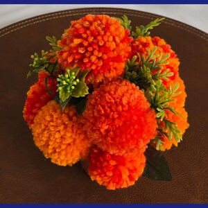 May include: A bouquet of orange and yellow yarn pom-poms arranged in a vase with green leaves. The pom-poms are arranged in a circular shape and resemble marigolds.