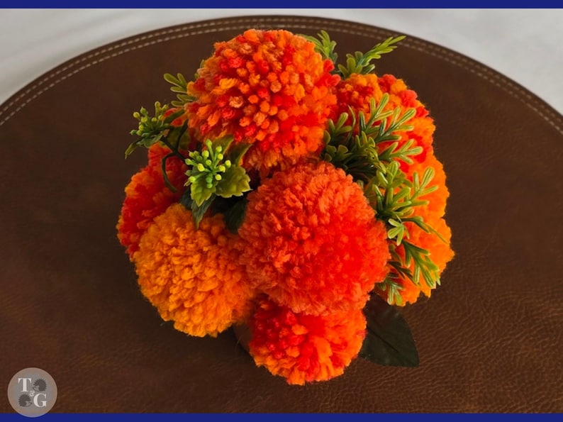 May include: A bouquet of orange and yellow yarn pom-poms arranged in a vase with green leaves. The pom-poms are arranged in a circular shape and resemble marigolds.