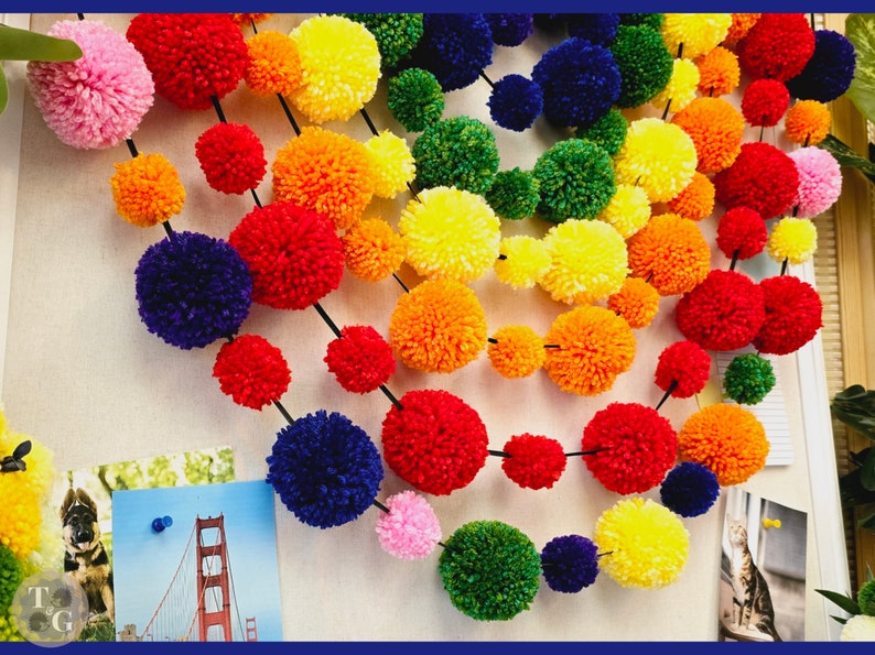 May include: A rainbow garland made of colorful yarn pom-poms. The garland is strung together with black thread and features a variety of colors, including red, orange, yellow, green, blue, and purple.