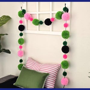 May include: A colorful pom-pom garland in green, pink, and black hangs in front of a white window frame. The garland is strung vertically and horizontally. Two pillows, one green and one striped, are below the garland.