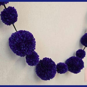 May include: A purple pom-pom garland with a black string connecting the pom-poms. The garland is hanging on a wall.