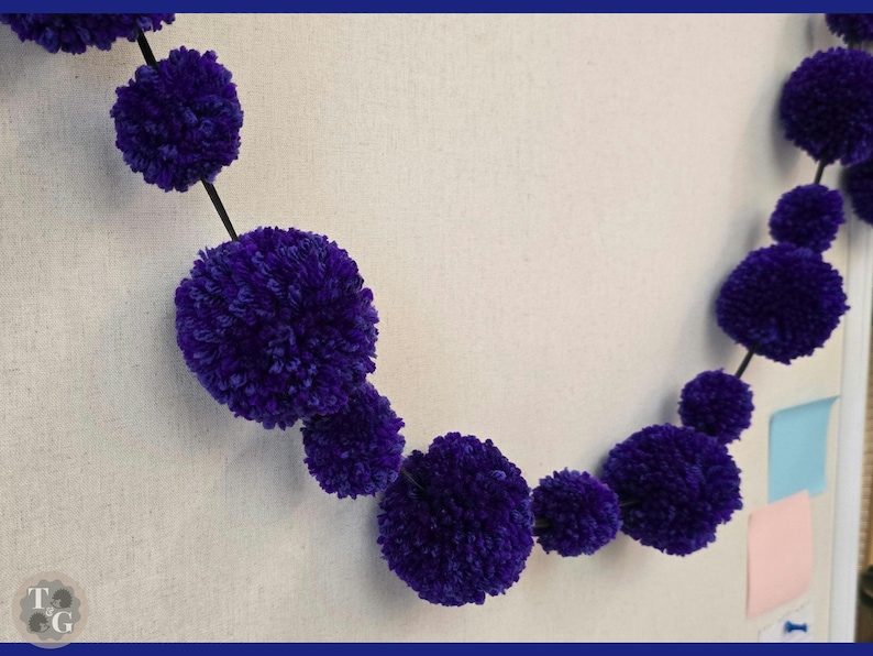 May include: A purple pom-pom garland with a black string connecting the pom-poms. The garland is hanging on a wall.