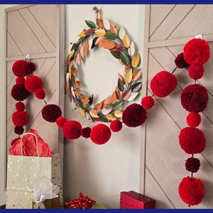 May include: A festive display featuring a fall-themed wreath and two strings of red pom-poms. The wreath is circular with colorful leaves. The pom-pom garlands are in varying shades of red. A gift bag and box are also present.