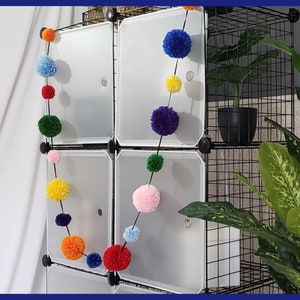 May include: A rainbow pom-pom garland hangs on a black wire shelf unit. The garland is made of colorful yarn pom-poms, including red, orange, yellow, green, blue, and pink. A potted plant sits on the shelf unit.