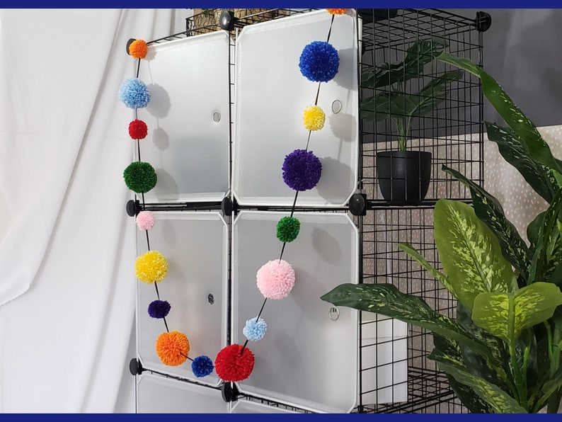 May include: A rainbow pom-pom garland hangs on a black wire shelf unit. The garland is made of colorful yarn pom-poms, including red, orange, yellow, green, blue, and pink. A potted plant sits on the shelf unit.