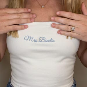 May include: A white tube top with the embroidered text "Mrs. Bowlin" in light blue. The top is paired with blue denim jeans. A gold necklace with a square pendant and a gold ring with a large square diamond are also visible.