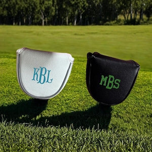 May include: Two golf club head covers on a green golf course. One is white with teal initials "KBL", the other is black with green initials "MBS". The covers are made of a leather-like material.