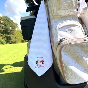 May include: White golf towel with a red golf cart design and the name "Katrina" embroidered in red script. The towel is hanging on a golf cart next to a golf bag with a floral pattern.