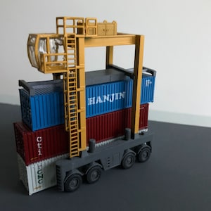 Peut inclure: Une grue jouet avec un cadre jaune et une base grise soulève une pile de quatre conteneurs d'expédition. Les conteneurs sont bleus, rouges et blancs avec le texte "HANJIN", "Citi" et "H.OZU" imprimé dessus.