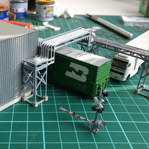 Puede incluir: Un set de tren modelo con un vagón verde, un camión blanco y un edificio gris. El set de tren está en una estera de corte verde con un cruce de ferrocarril.