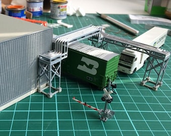 May include: A model train set with a green boxcar, a white semi-truck, and a gray building. The train set is on a green cutting mat with a railroad crossing.