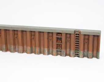 May include: A brown and gray model of a concrete seawall with a ladder on the right side. The wall has a textured surface and a gray concrete top.