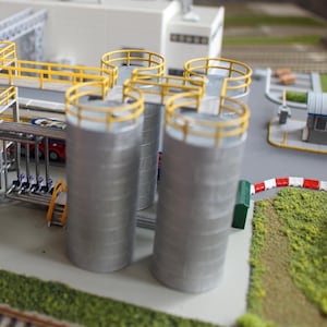 Puede incluir: Una maqueta de una instalación industrial que presenta cuatro tanques de almacenamiento cilíndricos de color plateado, rematados con barandillas de seguridad amarillas. Los tanques están conectados por pasarelas amarillas elevadas y estructuras de tuberías metálicas, situados en un paisaje en miniatura detallado con un edificio blanco.