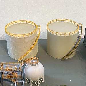May include: Two beige industrial storage tanks with yellow railings and staircases. The tanks are cylindrical with flat tops. Other industrial structures are visible in the background, suggesting a model or diorama of a factory or industrial site.