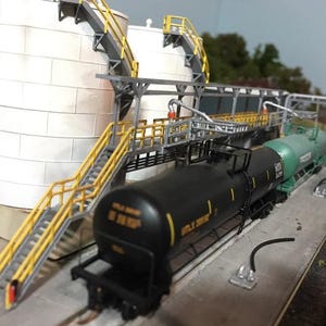 May include: A model train set with two black and green tanker cars, a white storage tank, and a metal framework with yellow handrails and stairs.