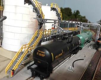 May include: A model train set with two black and green tanker cars, a white storage tank, and a metal framework with yellow handrails and stairs.