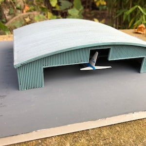 Può includere: Un modello di hangar per aerei in miniatura con un aereo blu e bianco all'interno. L'hangar è fatto di metallo ondulato grigio e ha un tetto curvo.