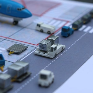 May include: A miniature airport scene with a blue airplane, white luggage carts, and white and blue vehicles. The scene is set on a gray surface with white lines.
