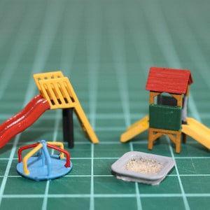 Può includere: Un set da gioco per parco giochi in miniatura con uno scivolo rosso e giallo, un giro-giostra blu e rosso, uno scivolo giallo e una casetta da gioco rossa e verde con una sabbiera.