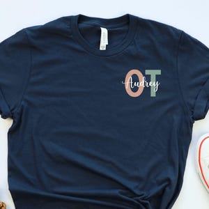May include: Navy blue t-shirt with the letters "OT" in peach and green, and the name "Audrey" in script. White sneakers with red trim are next to the shirt. Red berries and pinecones are on the left.