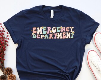 May include: A navy blue t-shirt with the text "EMERGENCY DEPARTMENT" in a retro font with colorful letters and small flower accents. The shirt is folded and tied in a knot.