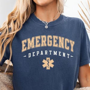 May include: A navy blue short-sleeve t-shirt featuring the text EMERGENCY DEPARTMENT in a curved beige font, with the word DEPARTMENT in a smaller straight font below, and a beige Star of Life medical symbol centered at the bottom.