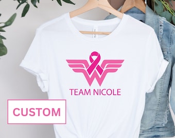 May include: White t-shirt with a pink Wonder Woman logo and a pink ribbon. The text "TEAM NICOLE" is printed below the logo. The word "CUSTOM" is printed in pink on a white background.
