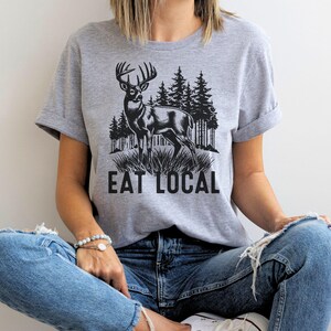 May include: A gray t-shirt with a black and white illustration of a deer in a forest setting. The text "Eat Local" is printed below the deer.