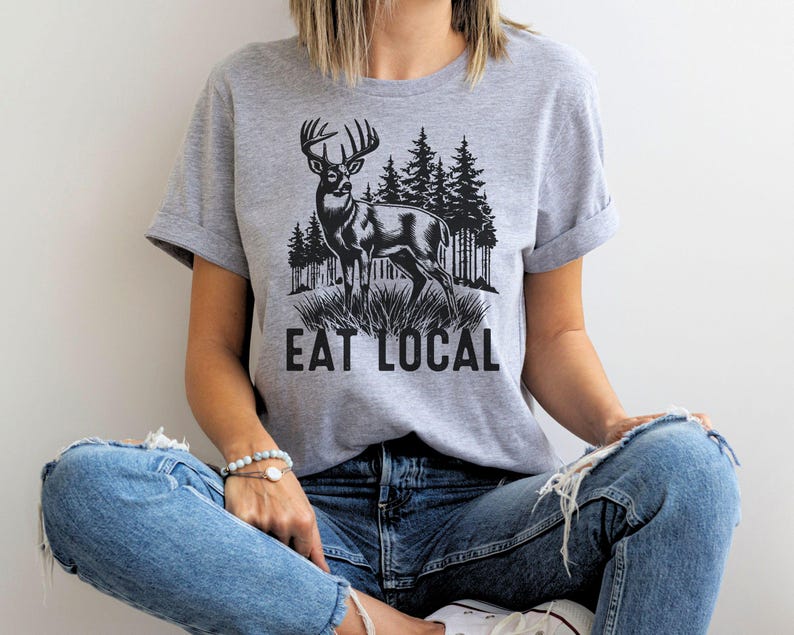 May include: A gray t-shirt with a black and white illustration of a deer in a forest setting. The text "Eat Local" is printed below the deer.