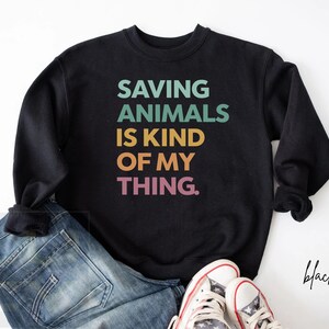 May include: Black sweatshirt with the text "SAVING ANIMALS IS KIND OF MY THING" in teal, green, yellow, and pink. The sweatshirt is paired with blue jeans and white sneakers.